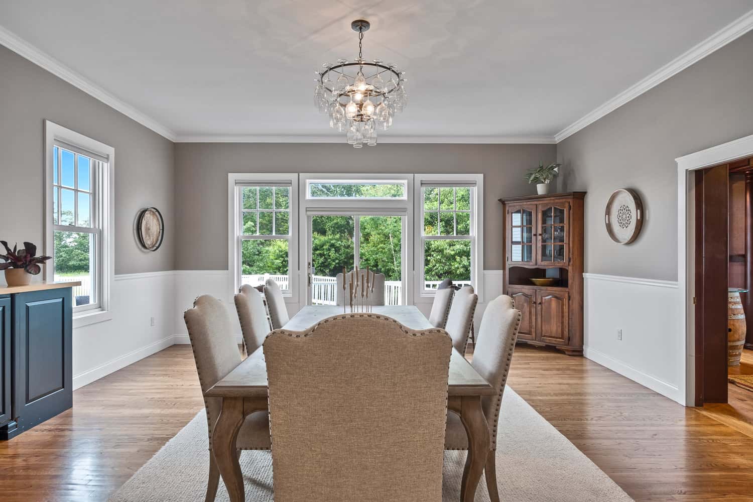 Elegant dining room with a wooden table, upholstered chairs, chandelier, and large windows with a view of greenery outside.