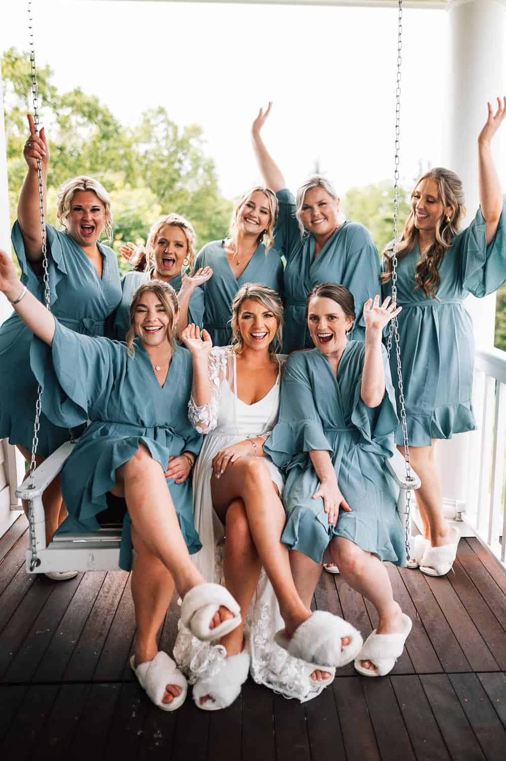 Bride and bridal party wearing slippers and sitting on a porch swing, smiling and waving.