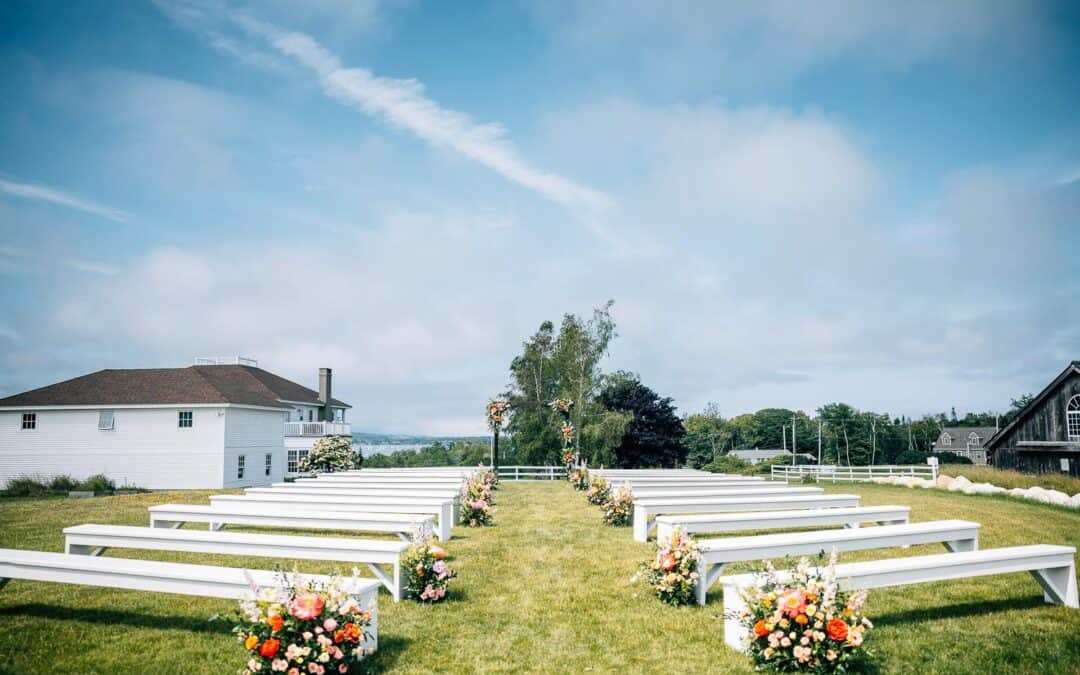 Luxury venues in coastal maine, outdoor wedding with rows of white benches adorned with colorful floral arrangements, set on a lush lawn under a blue sky,