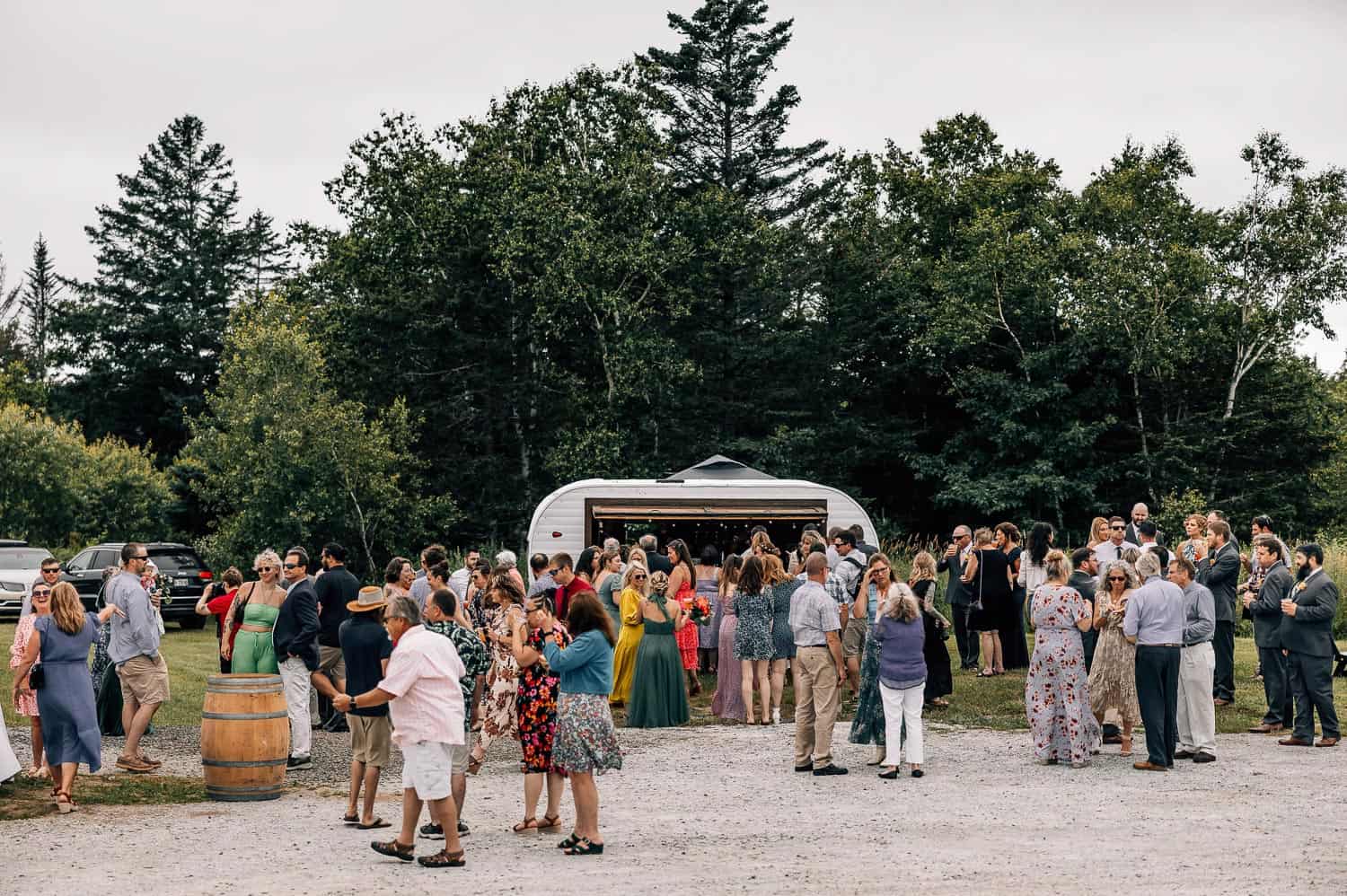 Outdoor gathering of people in semi-formal attire, mingling in a parking area near trees, with a white food truck and a barrel visible.