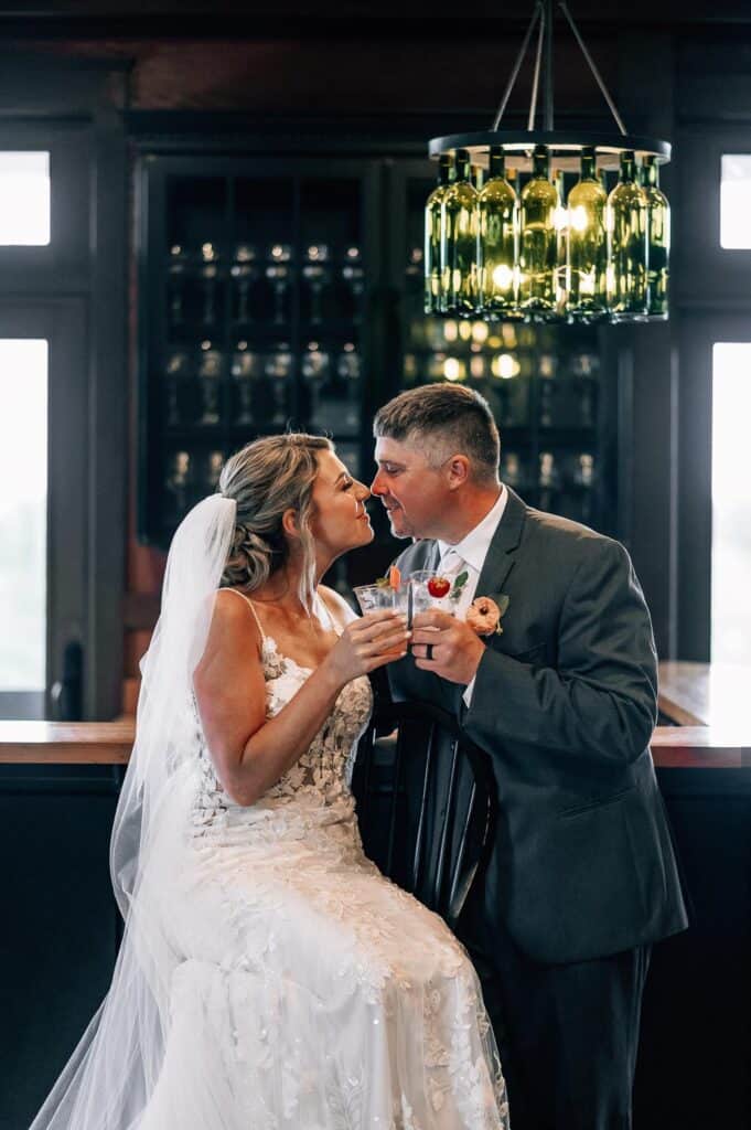 Rockland Maine Wedding Venues A bride and groom toast with drinks at a bar, with elegant lighting overhead and shelves of bottles in the background.