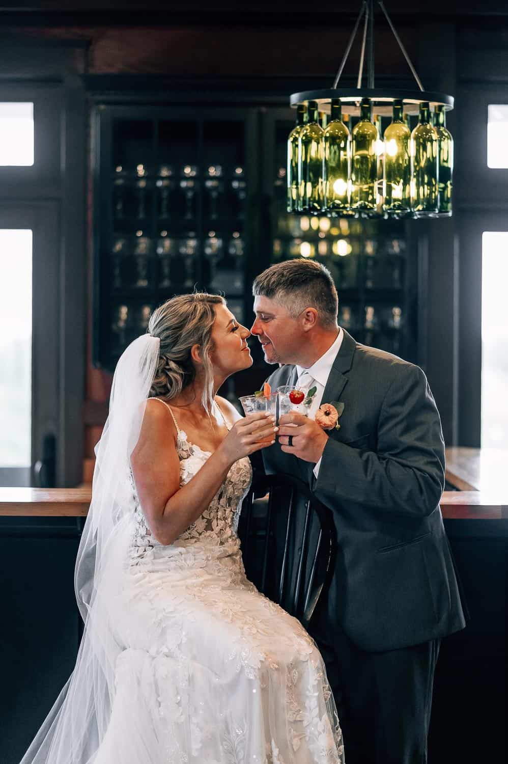 A bride and groom toast with drinks at a bar, with elegant lighting overhead and shelves of bottles in the background.