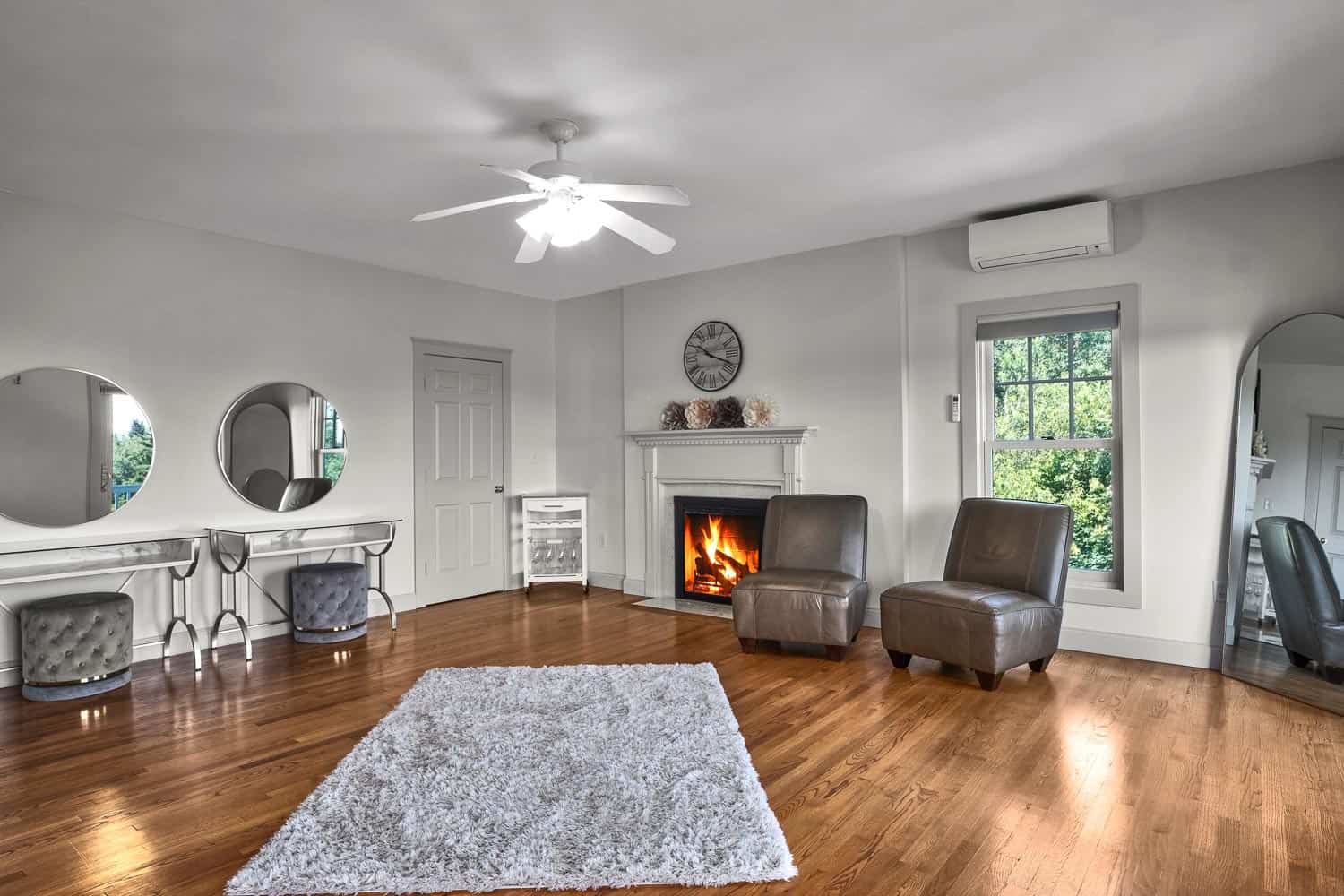 A spacious living room with a white rug, two grey armchairs, a lit fireplace, and multiple mirrors on the walls.