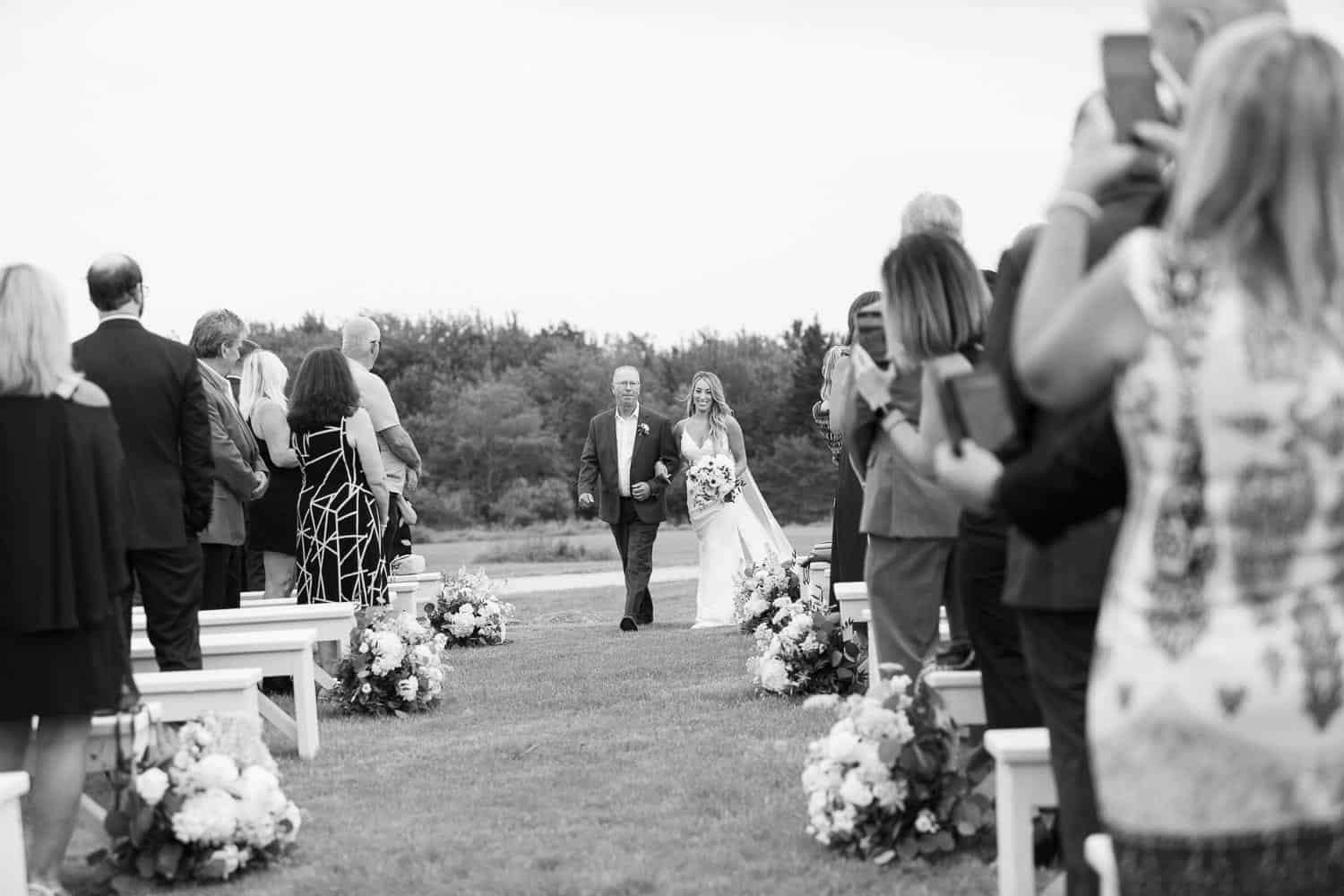 Black and white photo of guests watching a bride and her Dad walking down the aisle at an outdoor wedding ceremony.