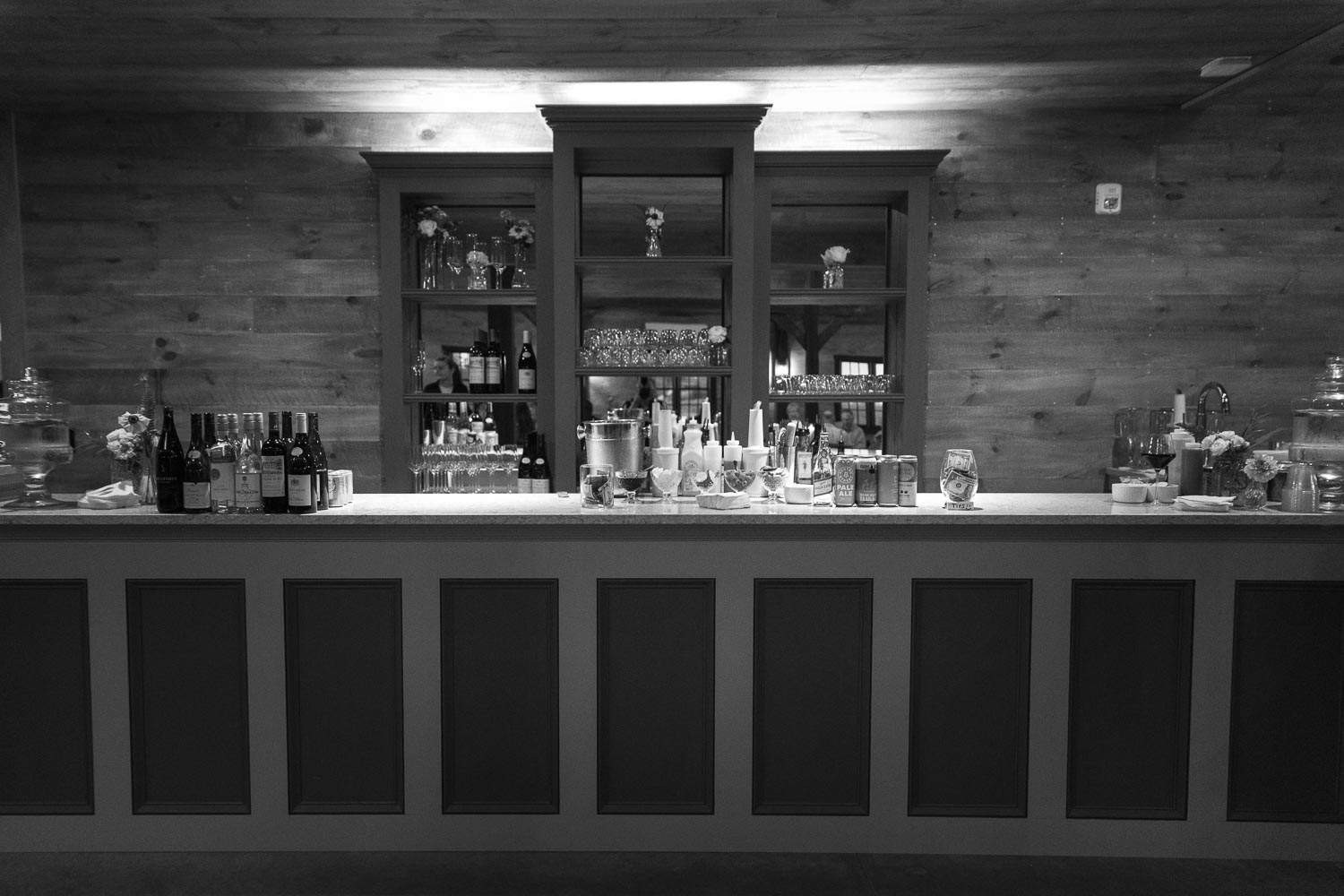 Black and white image of an empty bar counter with stools, bottles, and glasses, set against a wooden backdrop.