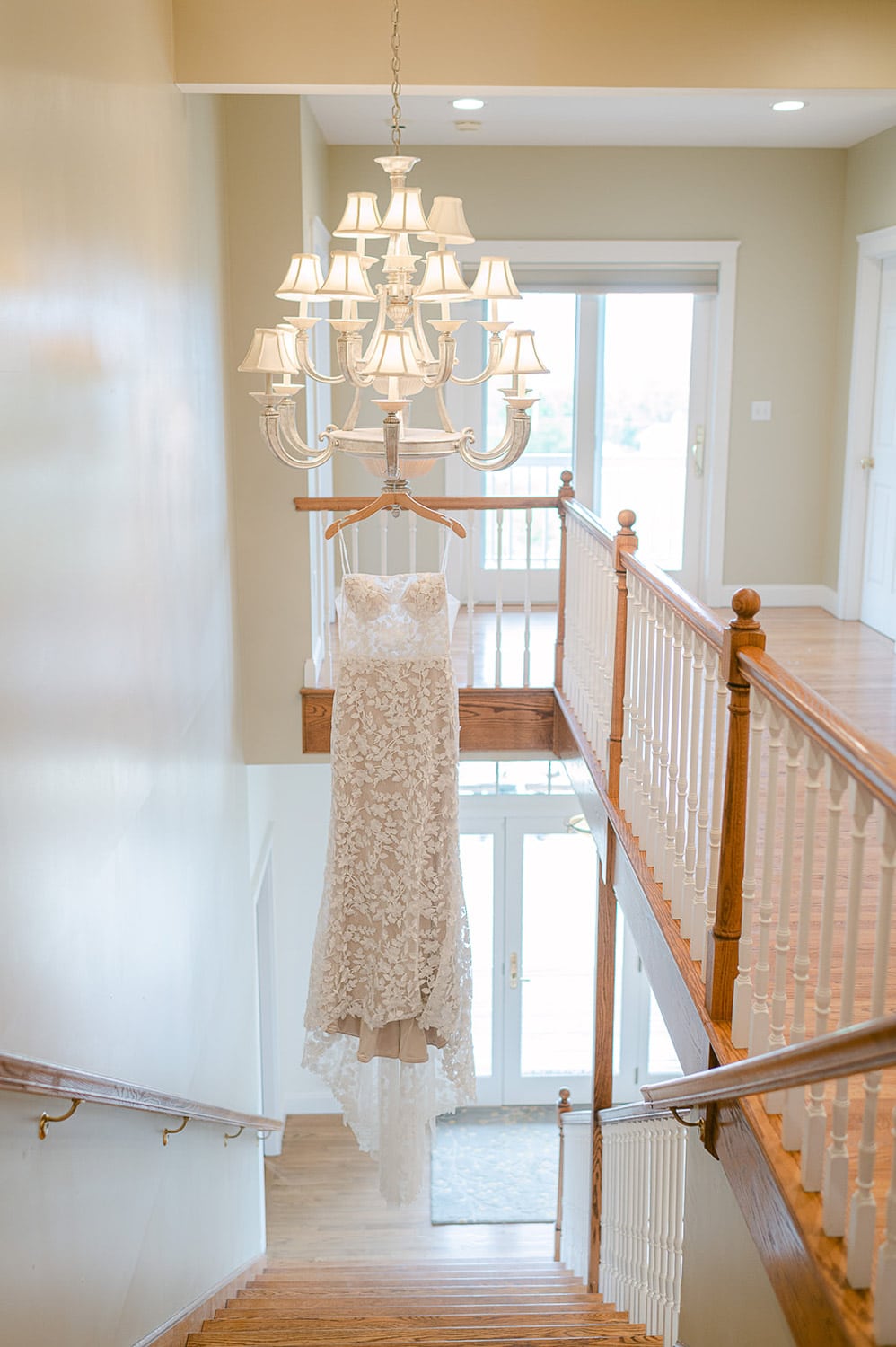 A lace wedding dress hangs from an elegant chandelier in a bright hallway with wooden stairs and a white railing.