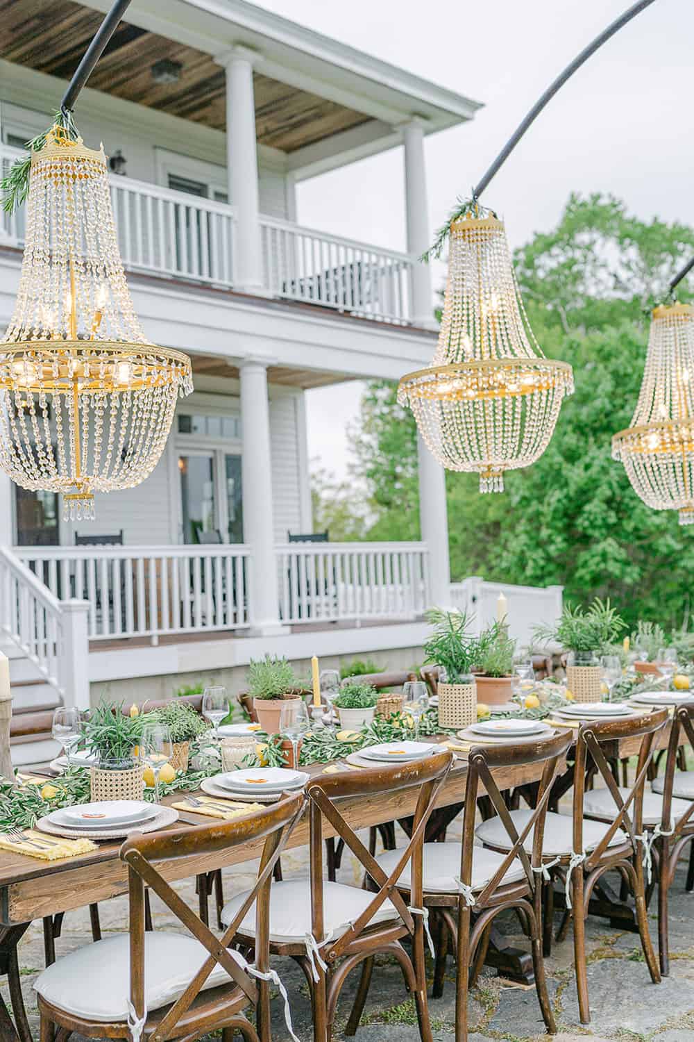 Elegant reception table with wooden chairs, adorned with greenery and chandeliers, in front of a white house with balconies.