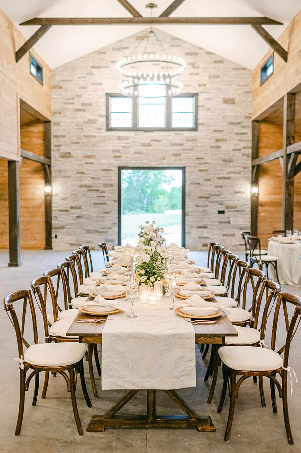 Elegant table inside a spacious hall with chairs, white dinnerware, and a central floral centerpiece, all under a rustic chandelier.