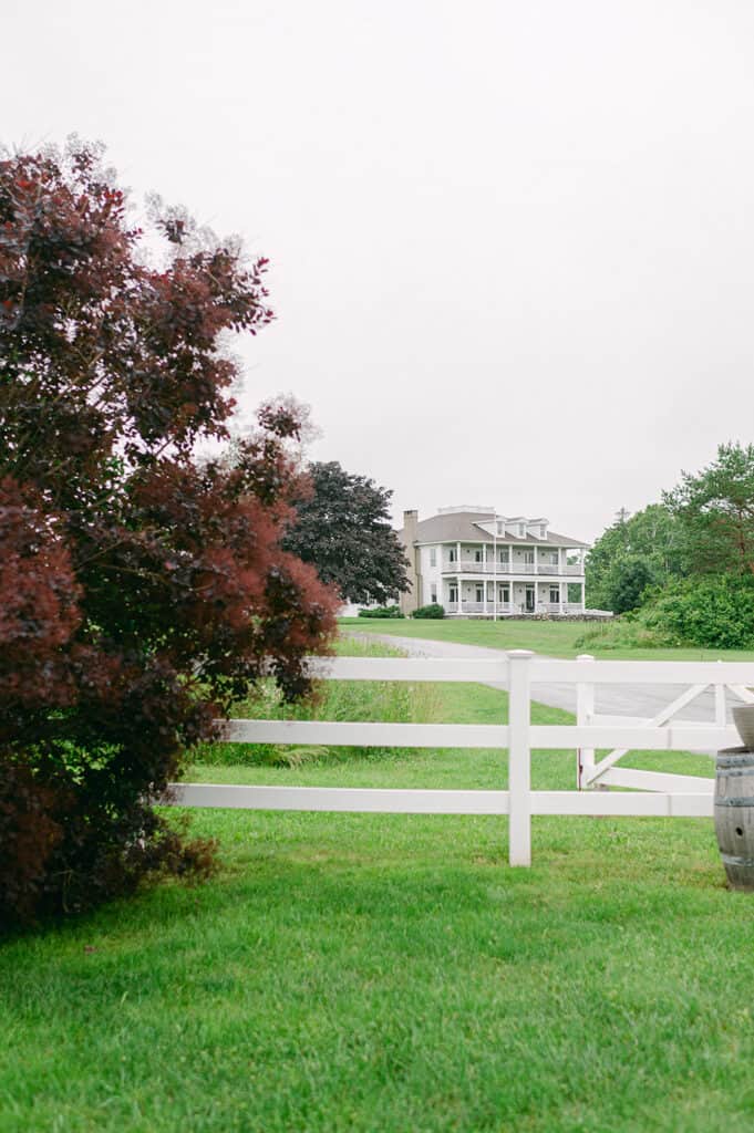 Rockland Maine Wedding Venues A white mansion at Ash Point Estate, viewed across a field with a white fence and a reddish-brown tree in the foreground.