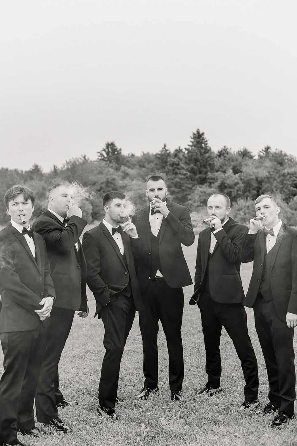 A group of six men in tuxedos, standing outdoors, each holding a cigar, with a natural backdrop.