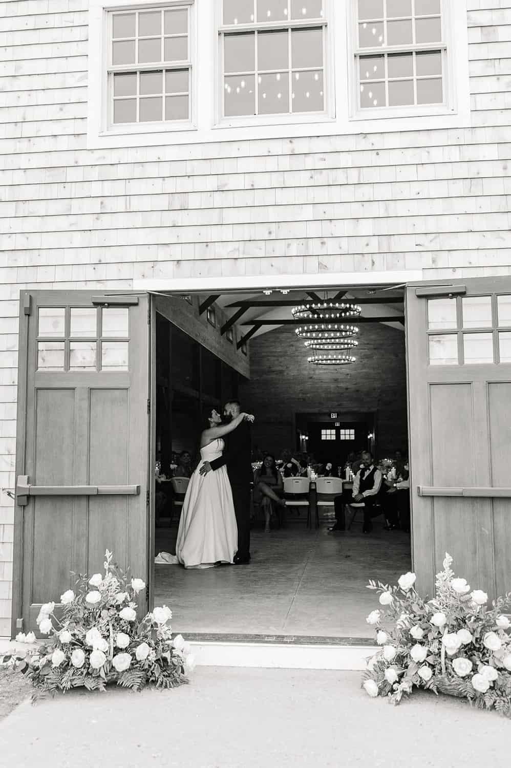 A black and white photo of a bride and groom dancing inside a barn with open doors, surrounded by guests and floral decorations.
