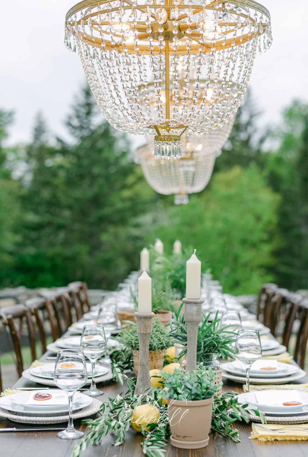 Elegant outdoor table with a crystal chandelier, adorned with candles, greenery, and set with fine dinnerware.