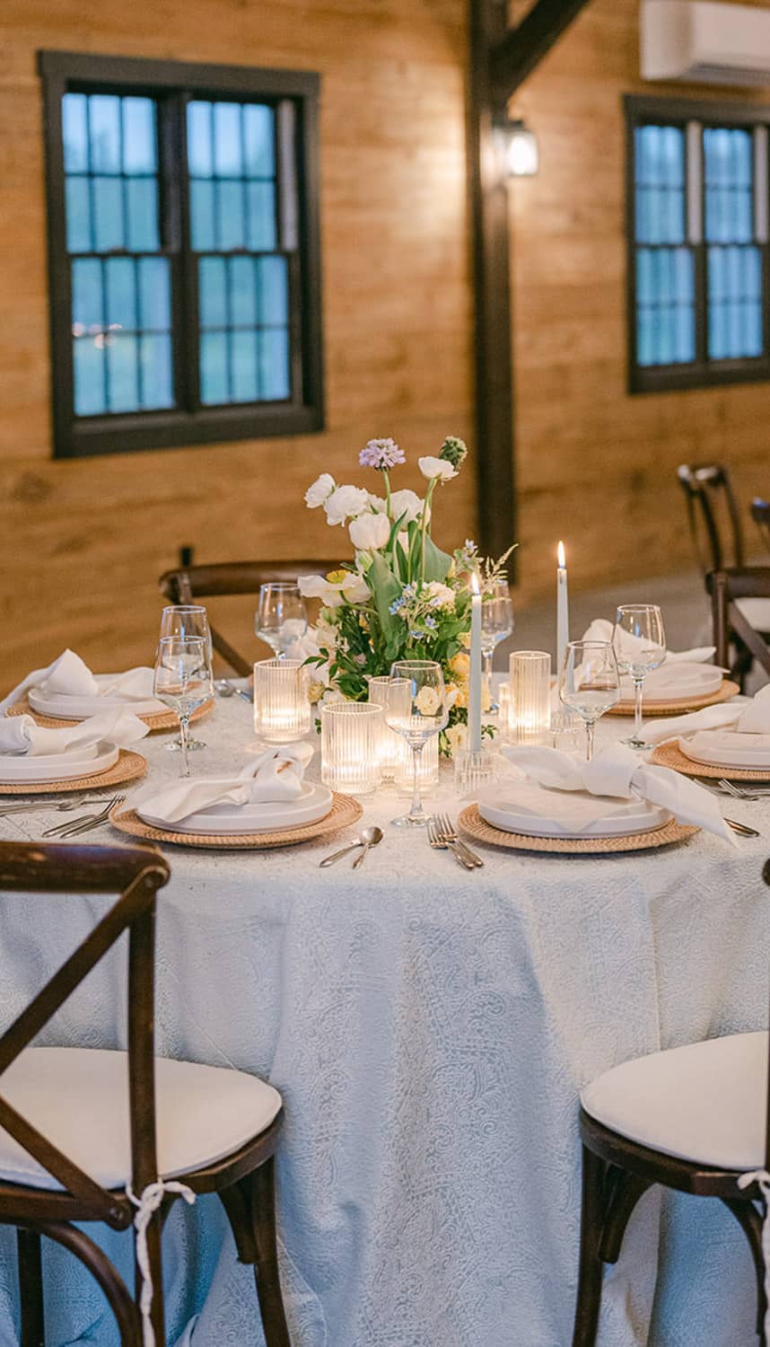 Elegant dining setup in a rustic venue with a floral centerpiece, candles, and a lace tablecloth, surrounded by wooden chairs.