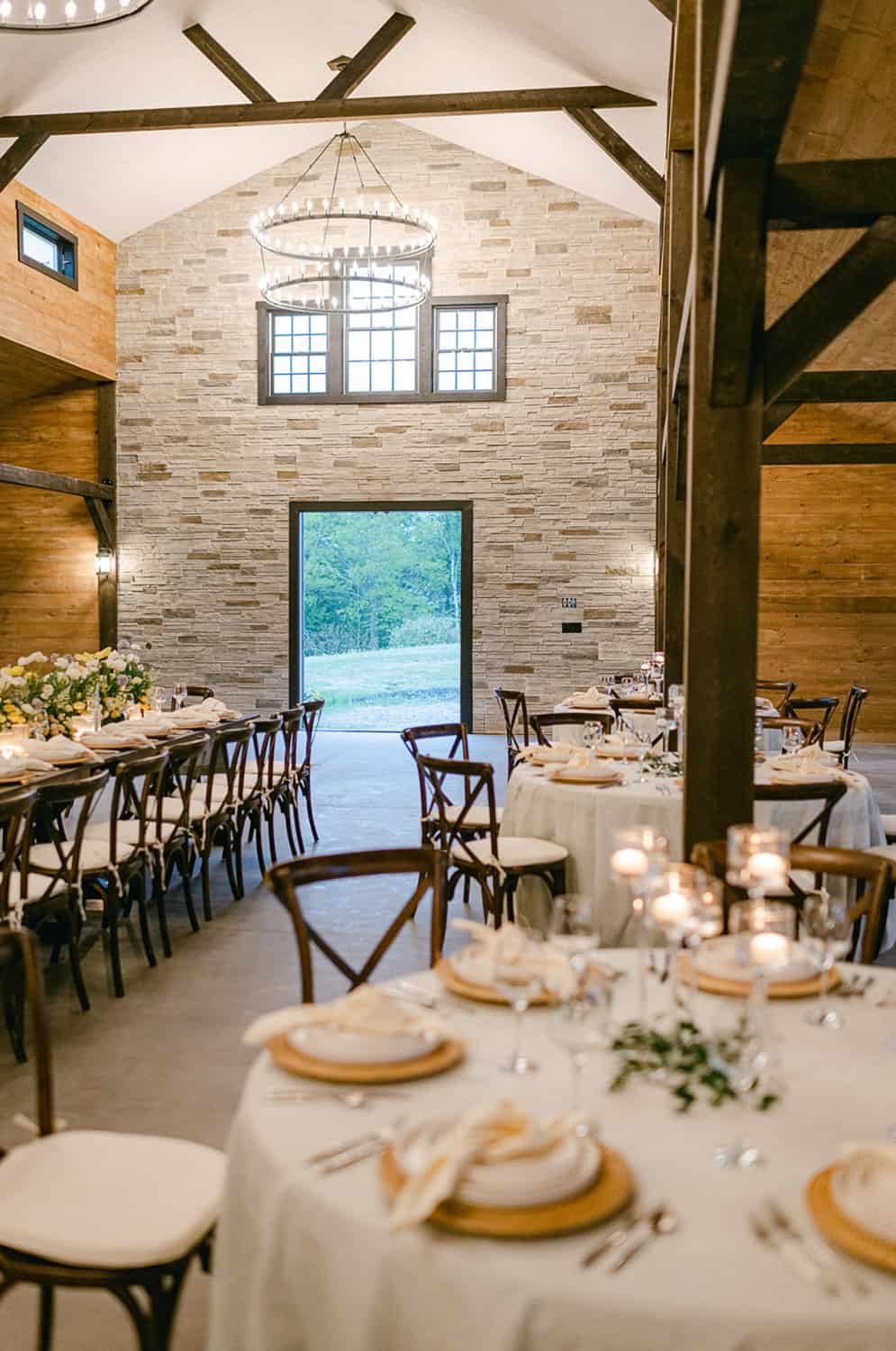 Rustic dining hall with round tables, white linen, wooden chairs, lit candles, stone walls, and wooden beams.