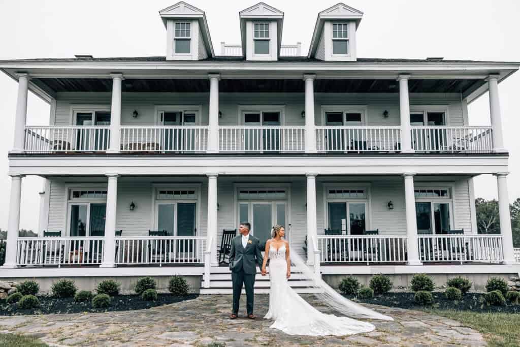 Maine coastal luxury wedding venues, a bride and groom holding hands in front of a large white house with double balconies, surrounded by a grassy lawn.