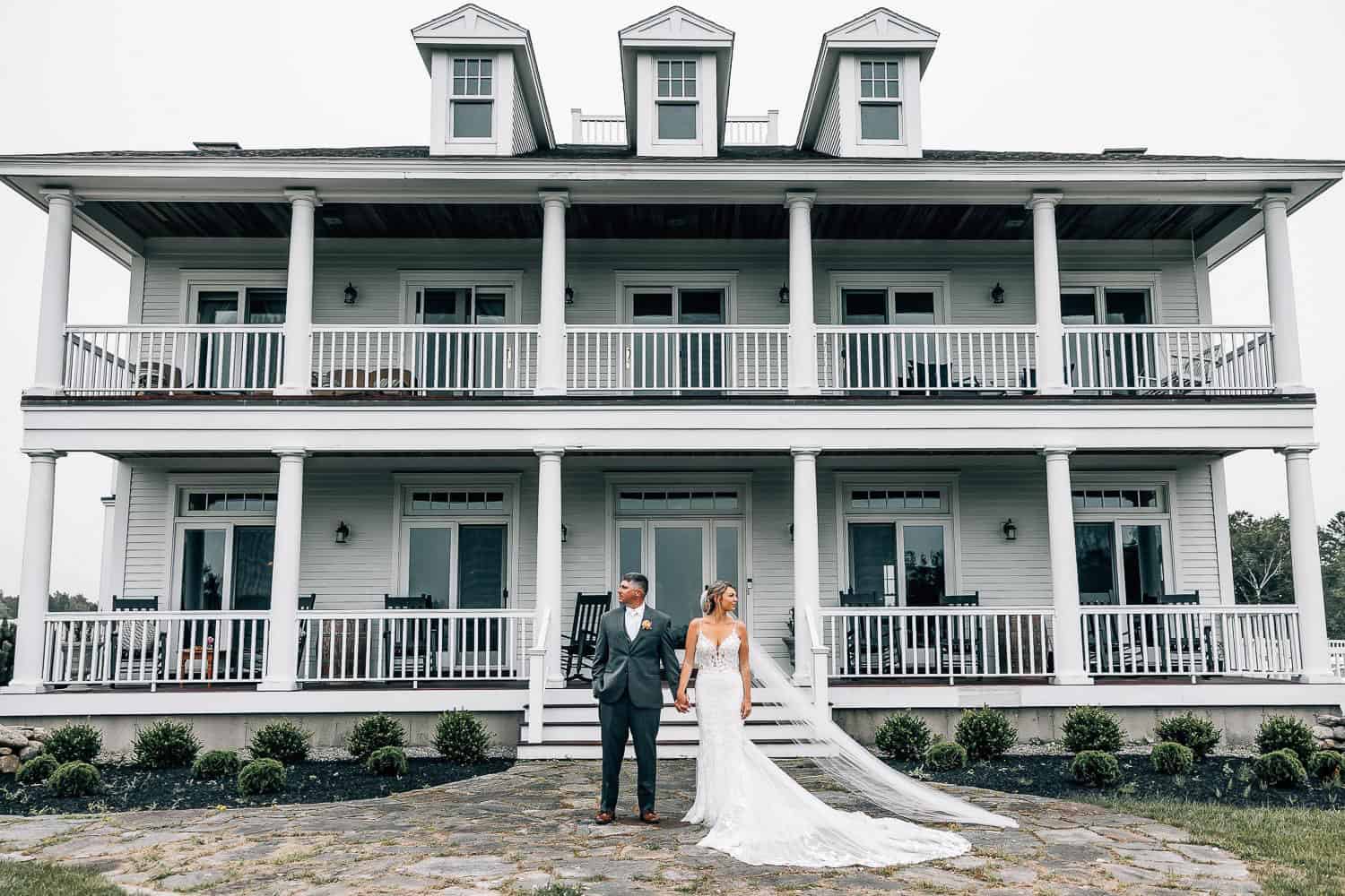 A bride and groom holding hands in front of a large white house with double balconies, surrounded by a grassy lawn.