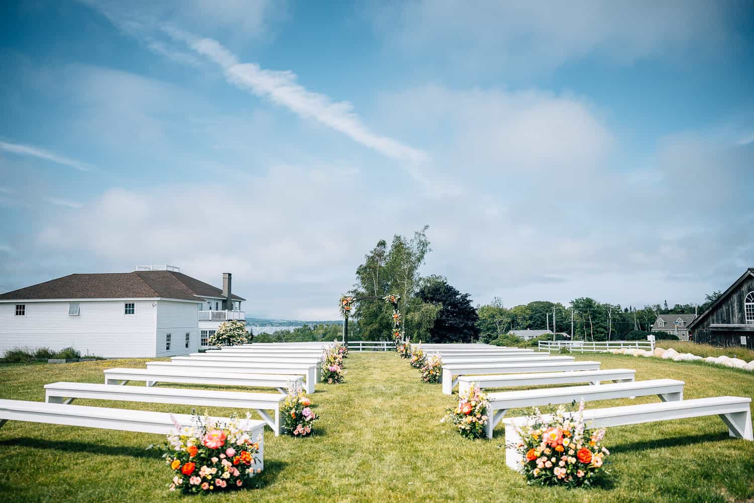 Outdoor wedding venue at Ash Point Estate, with white benches and floral decorations on a grassy field, under a blue sky with scattered clouds.