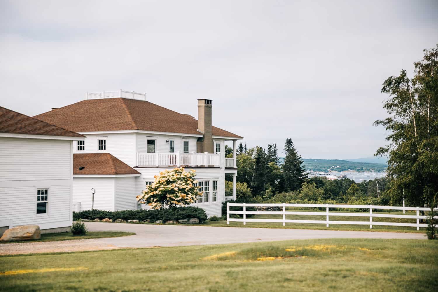 Large white house with multiple levels, chimney, and white fence, against a backdrop of grassy fields, and a distant harbor.