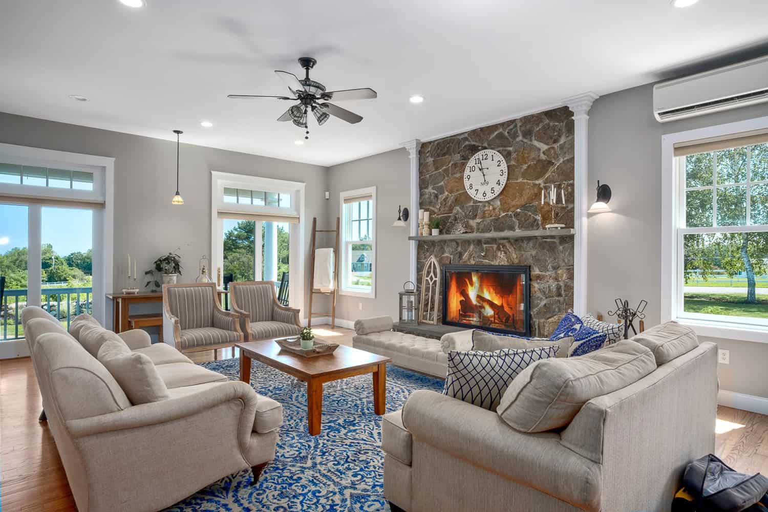 Spacious living room with stone fireplace, large windows, plush seating, and a blue area rug under a wooden coffee table.