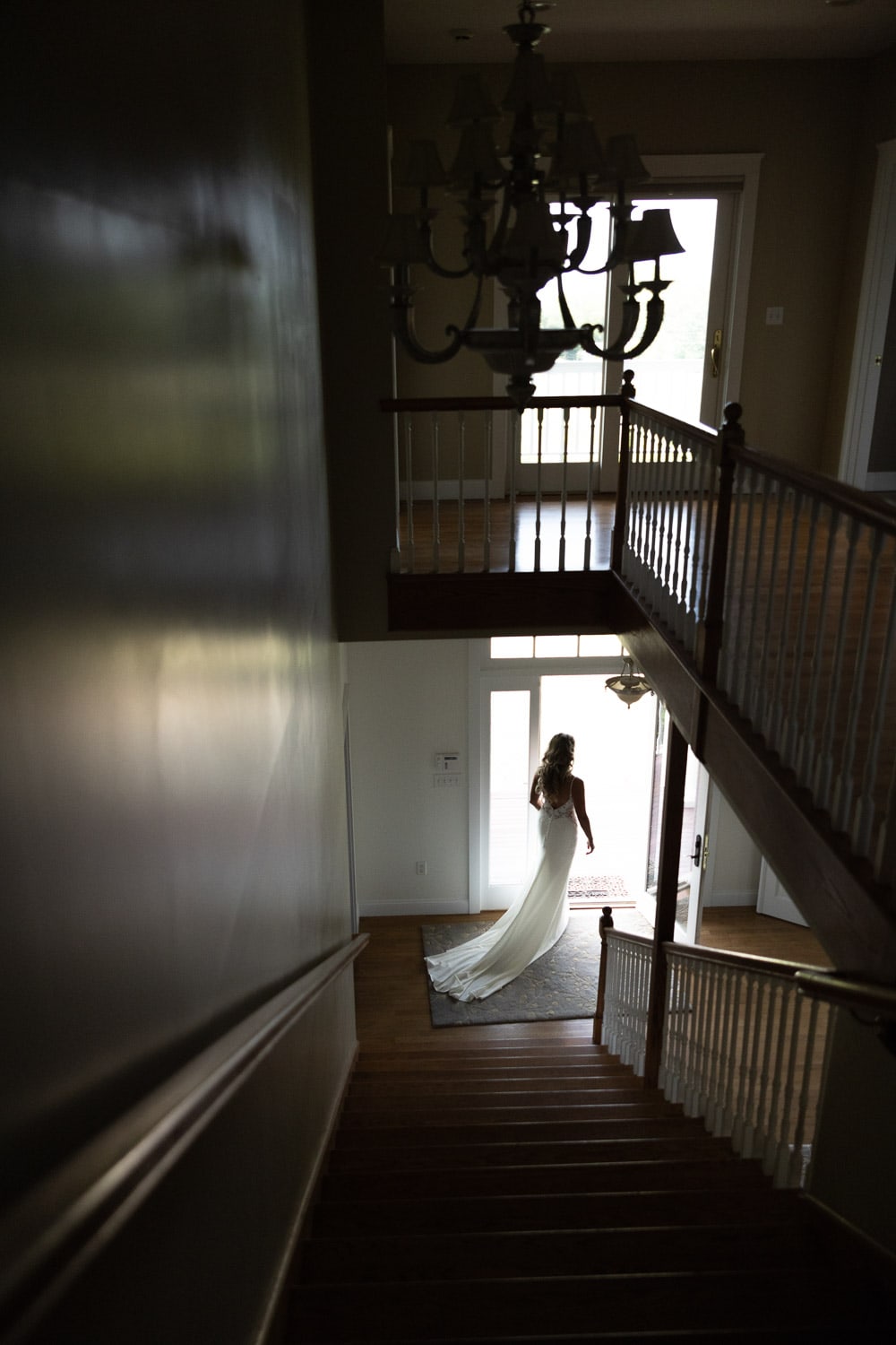 A bride in a long gown and veil descends a wooden staircase, viewed from the top of the stairs.