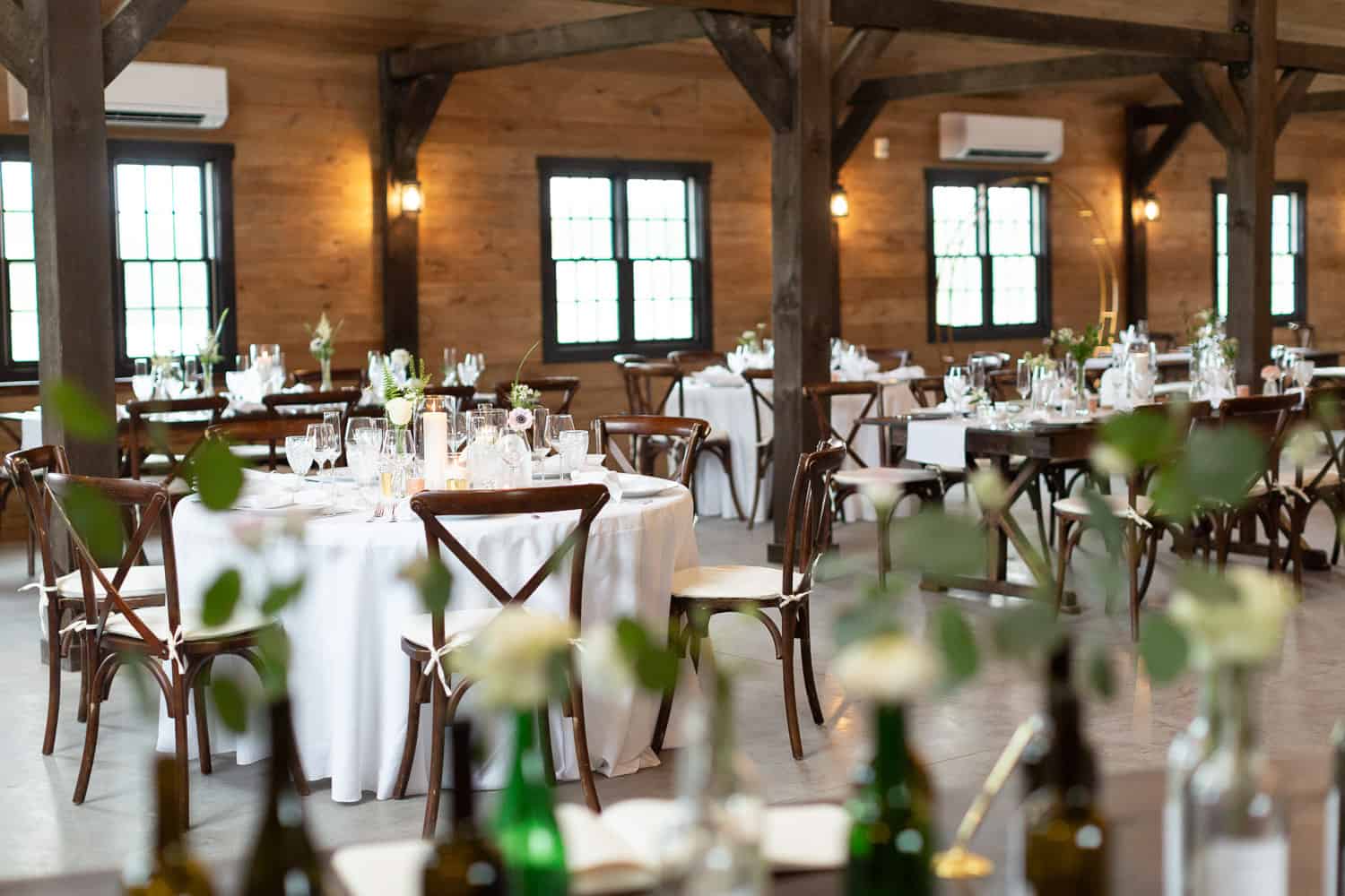 Ash Point Estate's elegant banquet hall: round tables, white linens, candles, wine bottles, exposed wooden beams, and large windows.