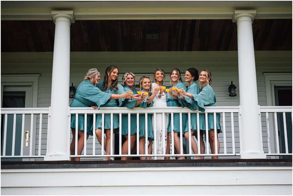 Luxury venues in coastal maine, a bride and her bridesmaids in matching robes have a toast on a porch with white columns.