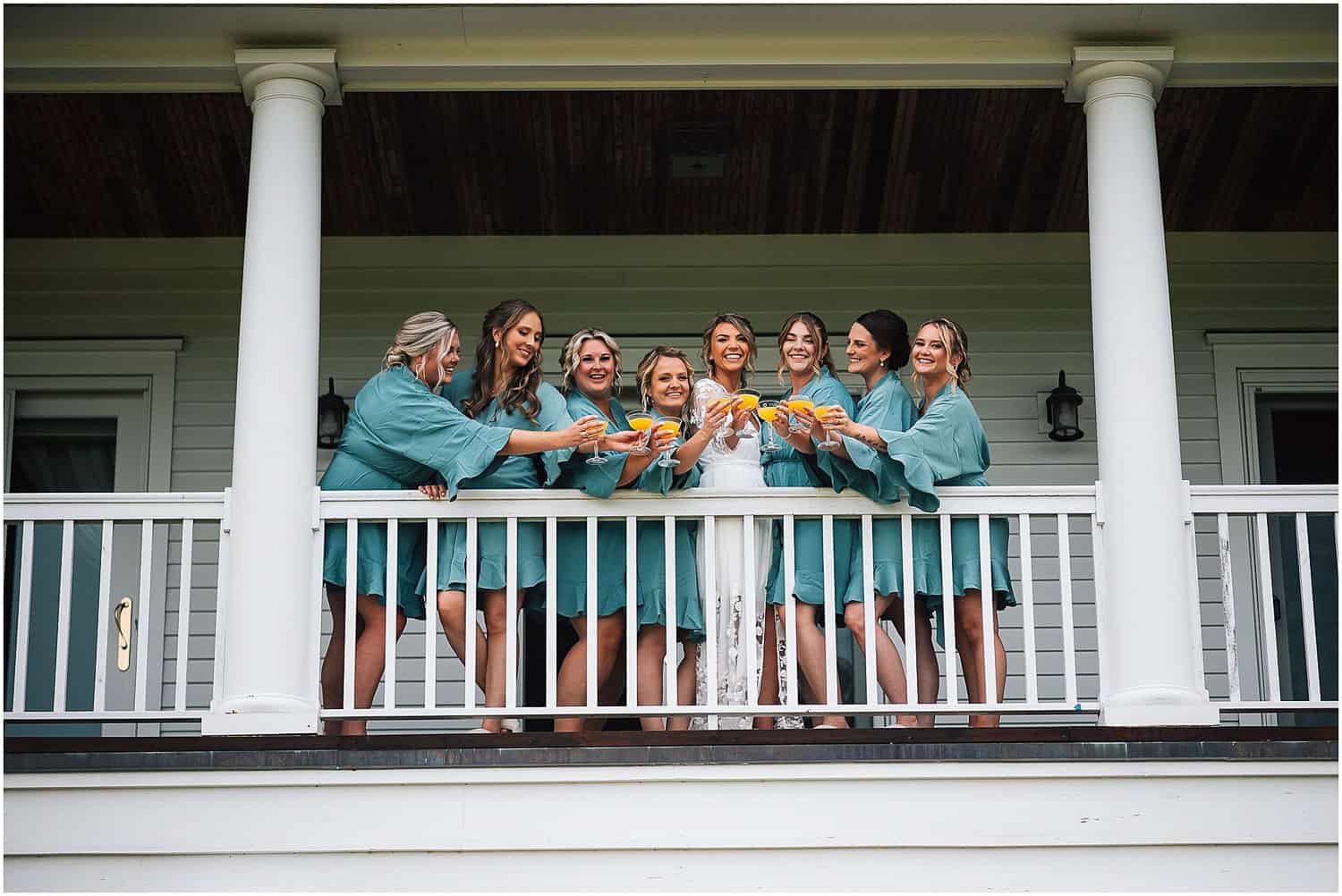 A bride and her bridesmaids in matching robes have a toast on a porch with white columns.