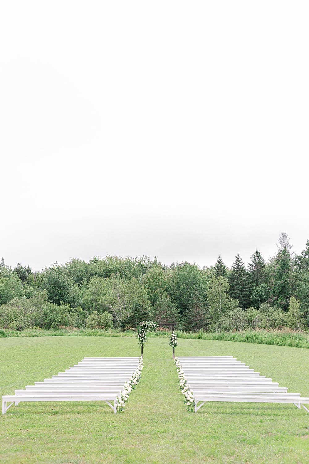 A set up for a wedding ceremony in a field with an arch and rows of white benches.