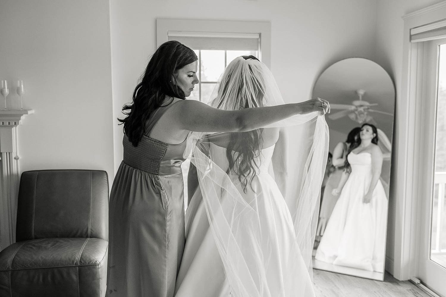 A woman adjusts the veil of a bride in front of a mirror in a softly lit room.
