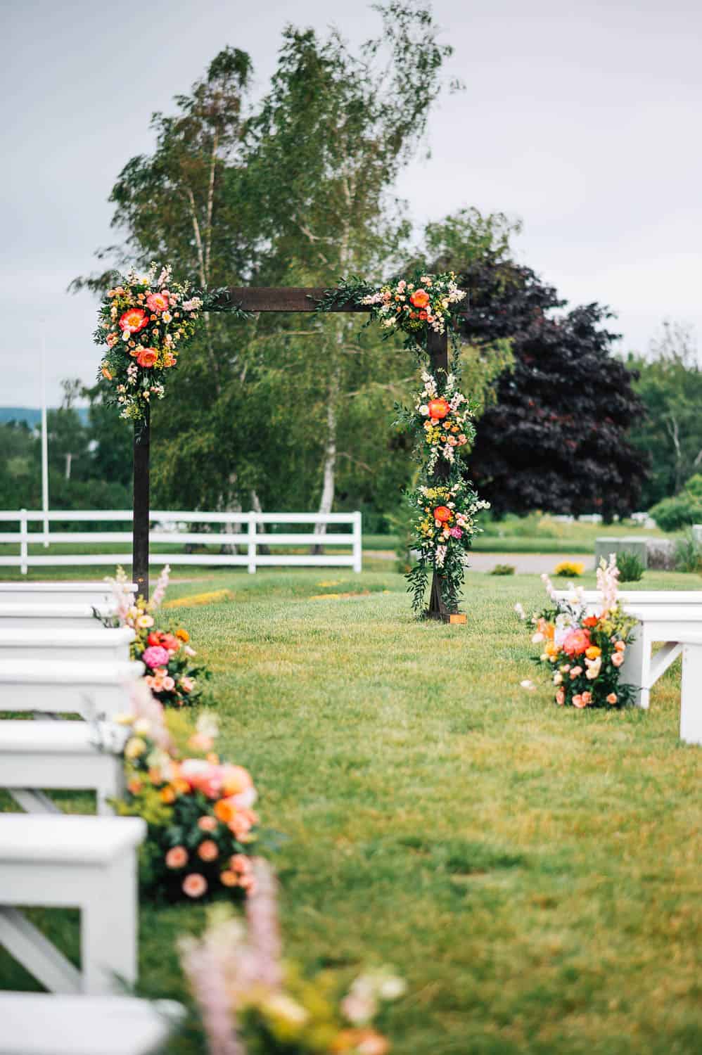 A wedding arch with flower bouquets on the ground at the end of white benches.