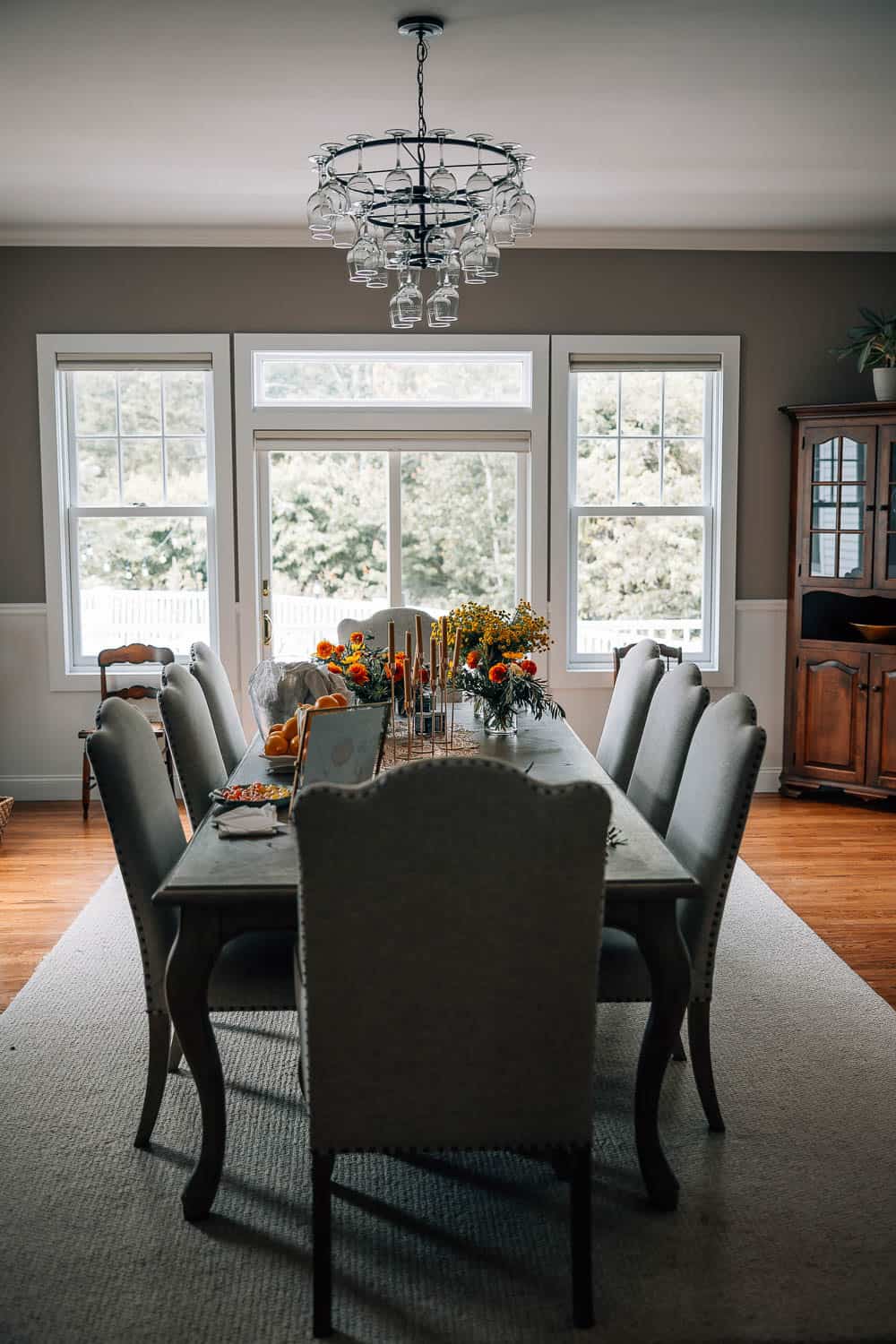 A dining room with a large table set for a meal, flanked by upholstered chairs and lit by a modern chandelier, with a view of trees through surrounding windows.