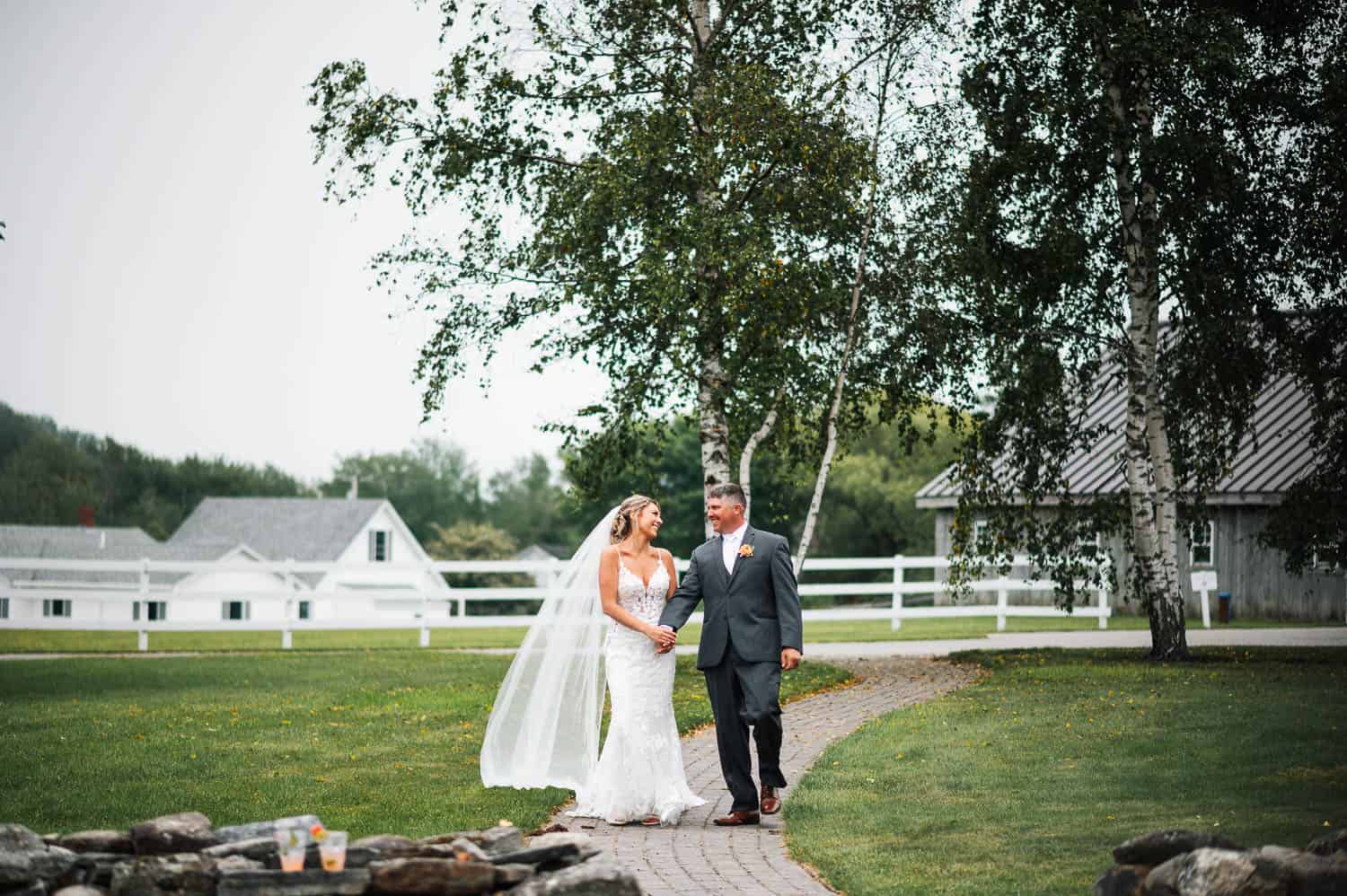A bride and groom walk hand in hand along a path, with trees and a white fence in the background, at a countryside setting.