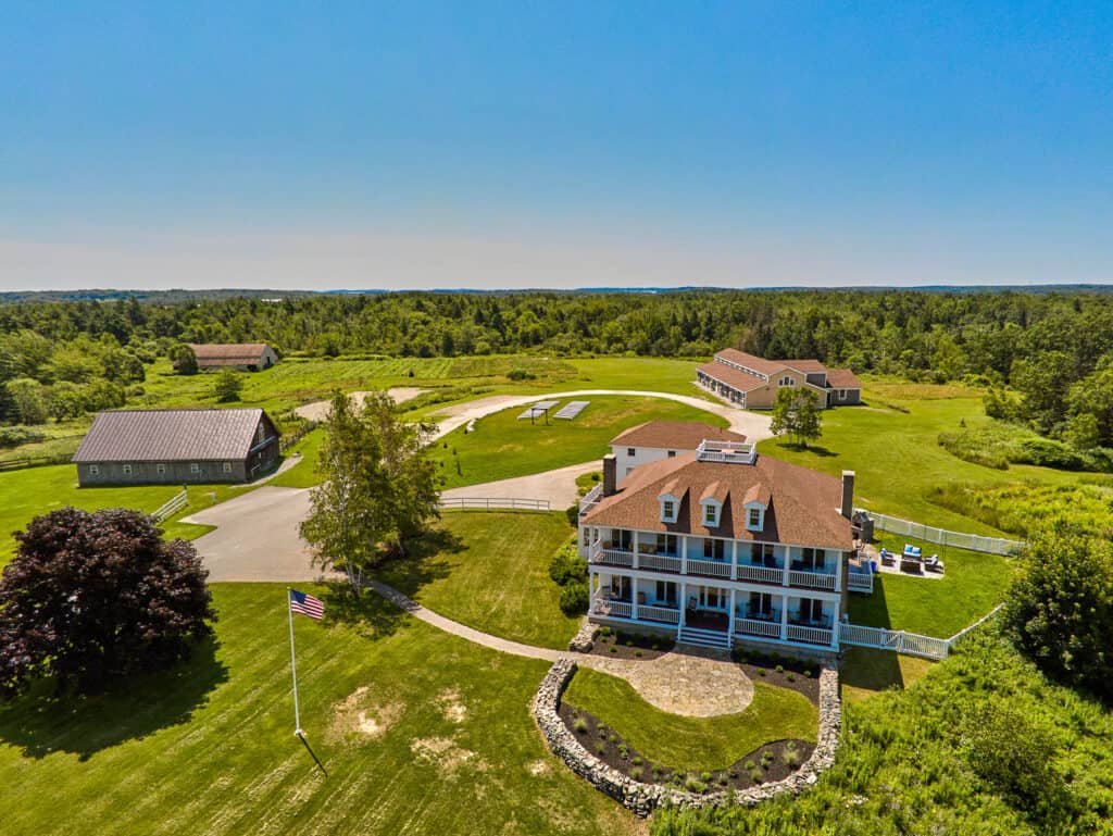 Private estate weddings in maine aerial view of ash point estate with a mansion, outbuildings, and landscaped lawns, surrounded by green fields under a clear blue sky.