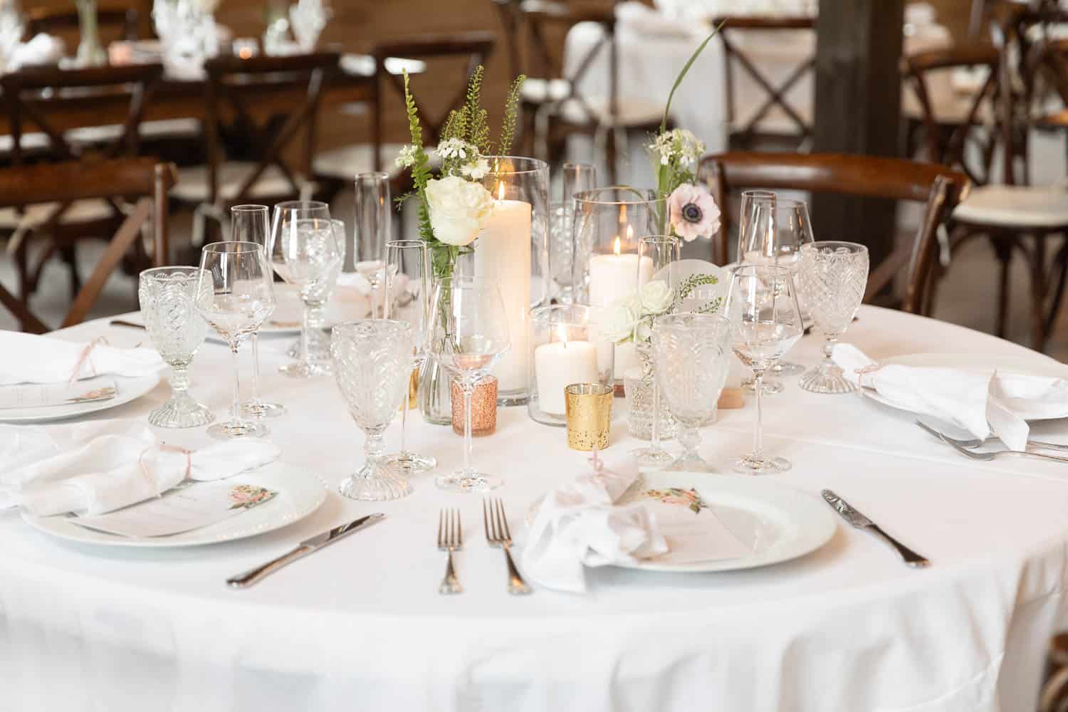 Elegant wedding table setting with crystal glassware, floral centerpieces, candles, and white linens in a well-lit venue.