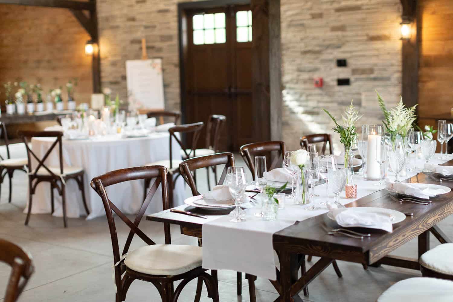 Elegant indoor dining setup at a wedding venue with white tablecloths, wooden chairs, and simple floral centerpieces.