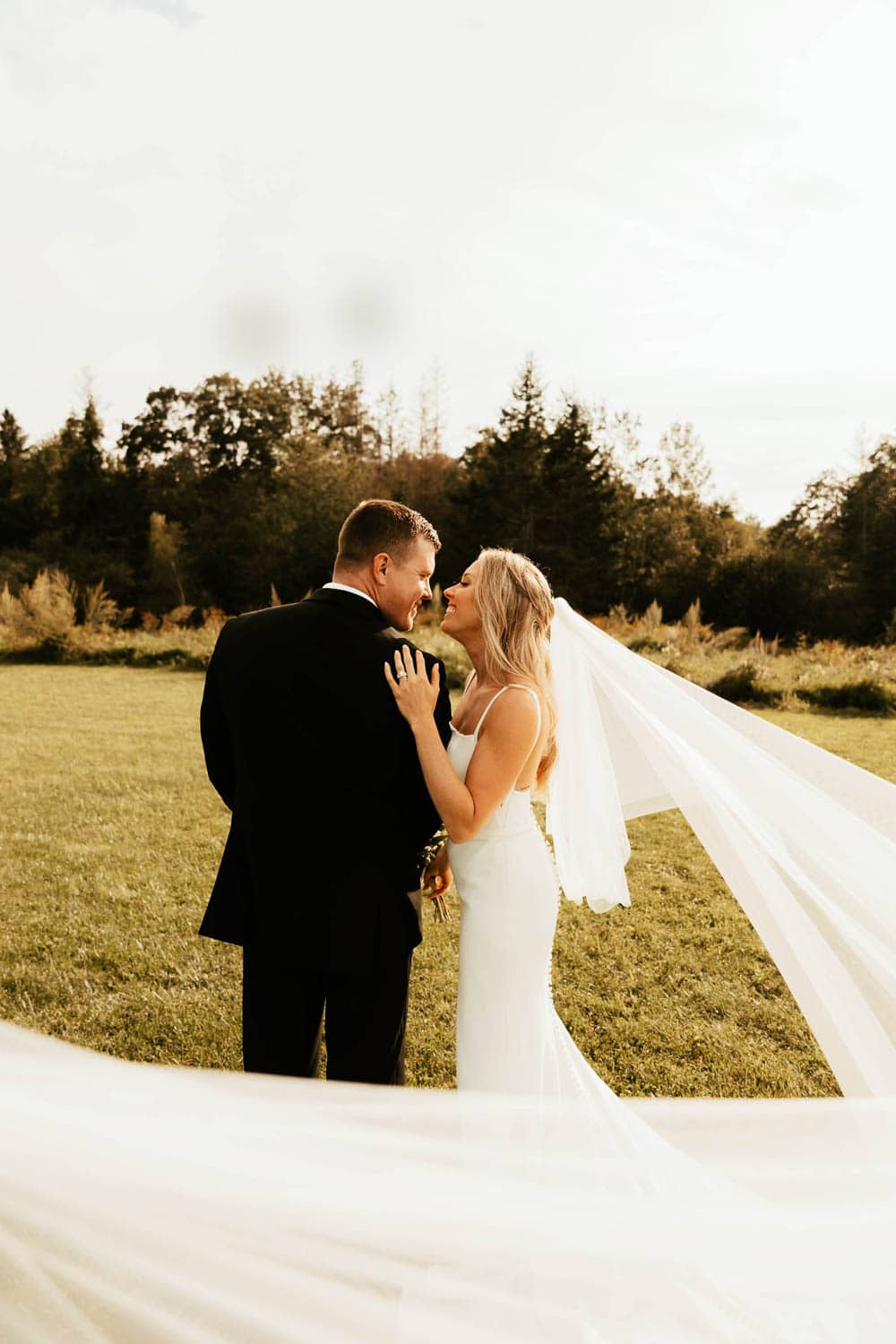 Bride with a flowing veil standing with her groom in a field.