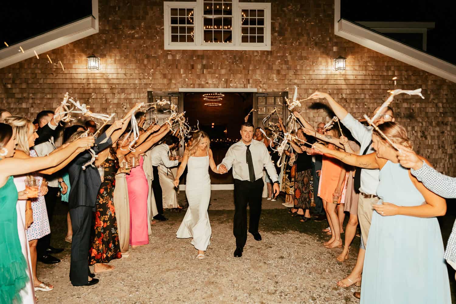 A bride and groom walking down a path between their guests who are waving balloons.