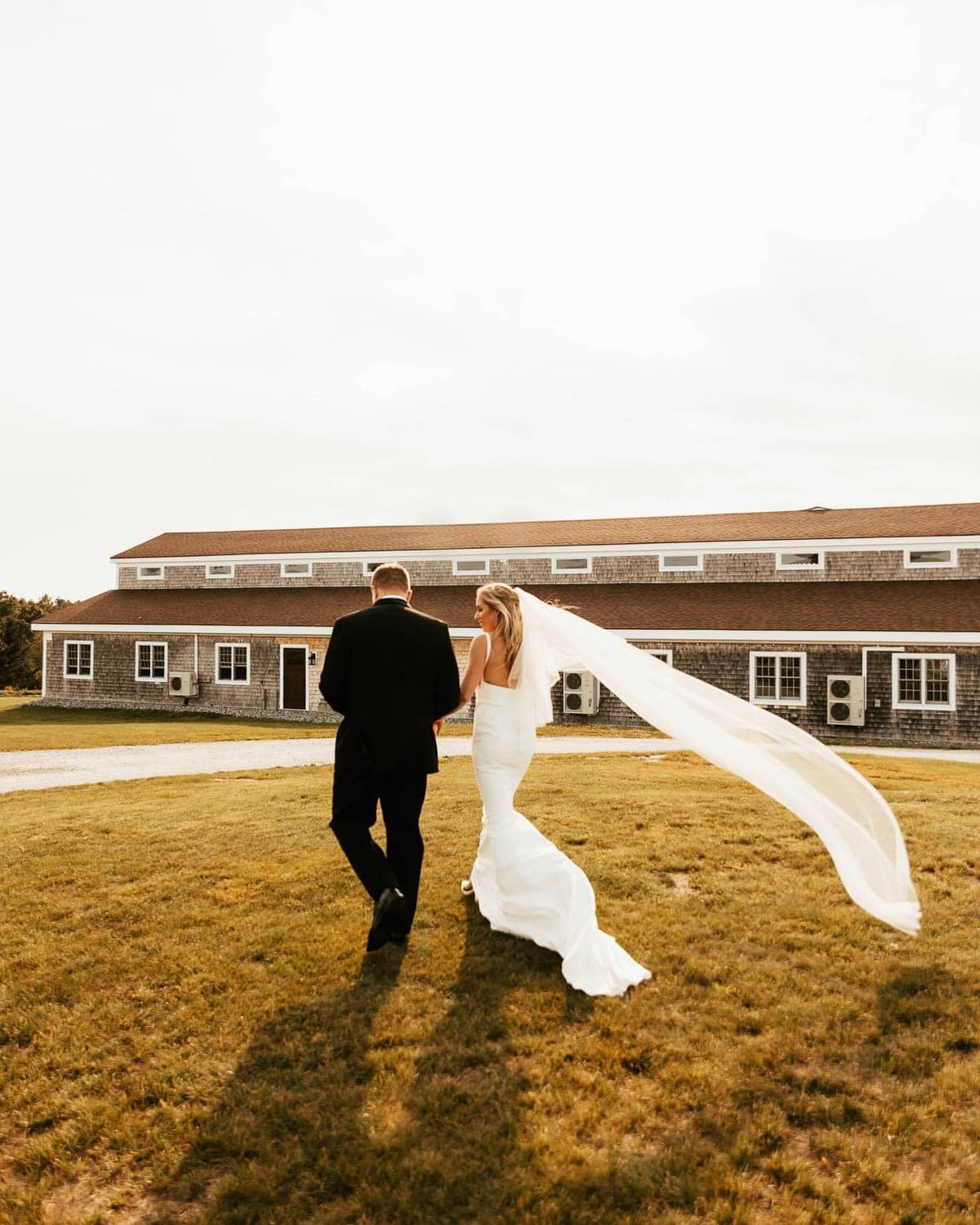 A bride with her veil trailing behind in the breeze, walking with her groom.