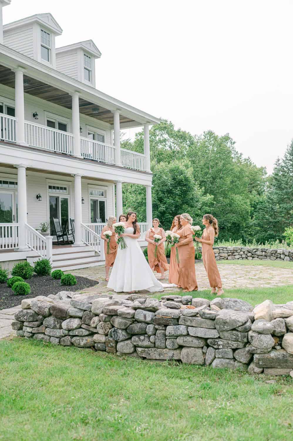 A bride and her bridesmaids holding bouquets standing on the lawn outside of a white mansion.
