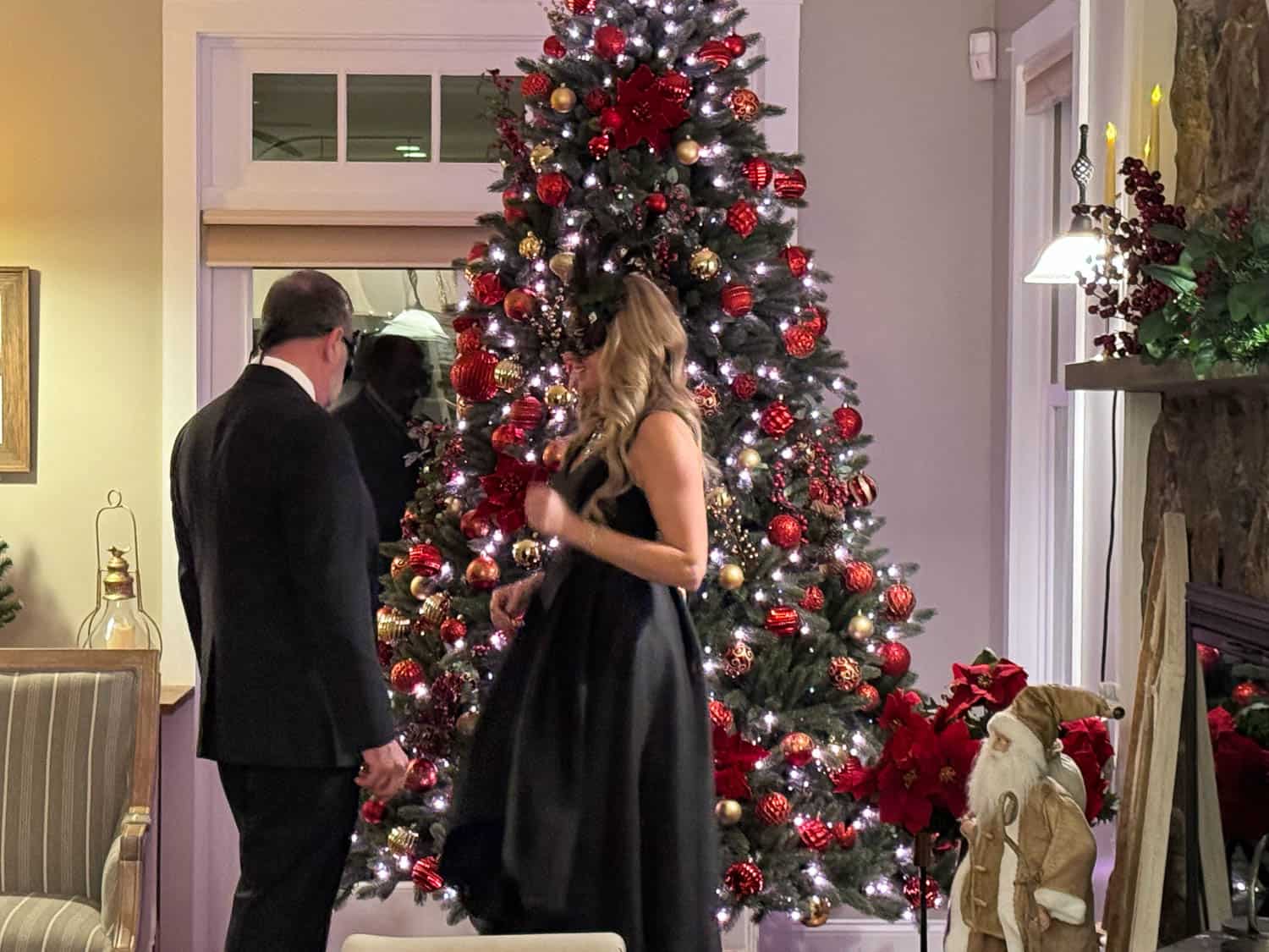 Two people in formal attire decorating a large, ornately decorated christmas tree in a warmly lit room.