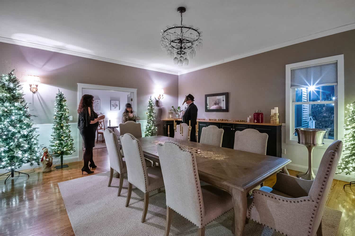 Interior of a dining room at Ash Point Estate, decorated for Christmas with multiple trees, featuring three people talking.