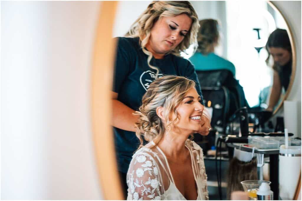 Estate Wedding Packages In Maine: What’s Included In A Luxury Experience 2 Luxury coastal maine wedding venues a bride smiling in a salon chair as a stylist fixes her hair, with other people and salon equipment blurred in the background.