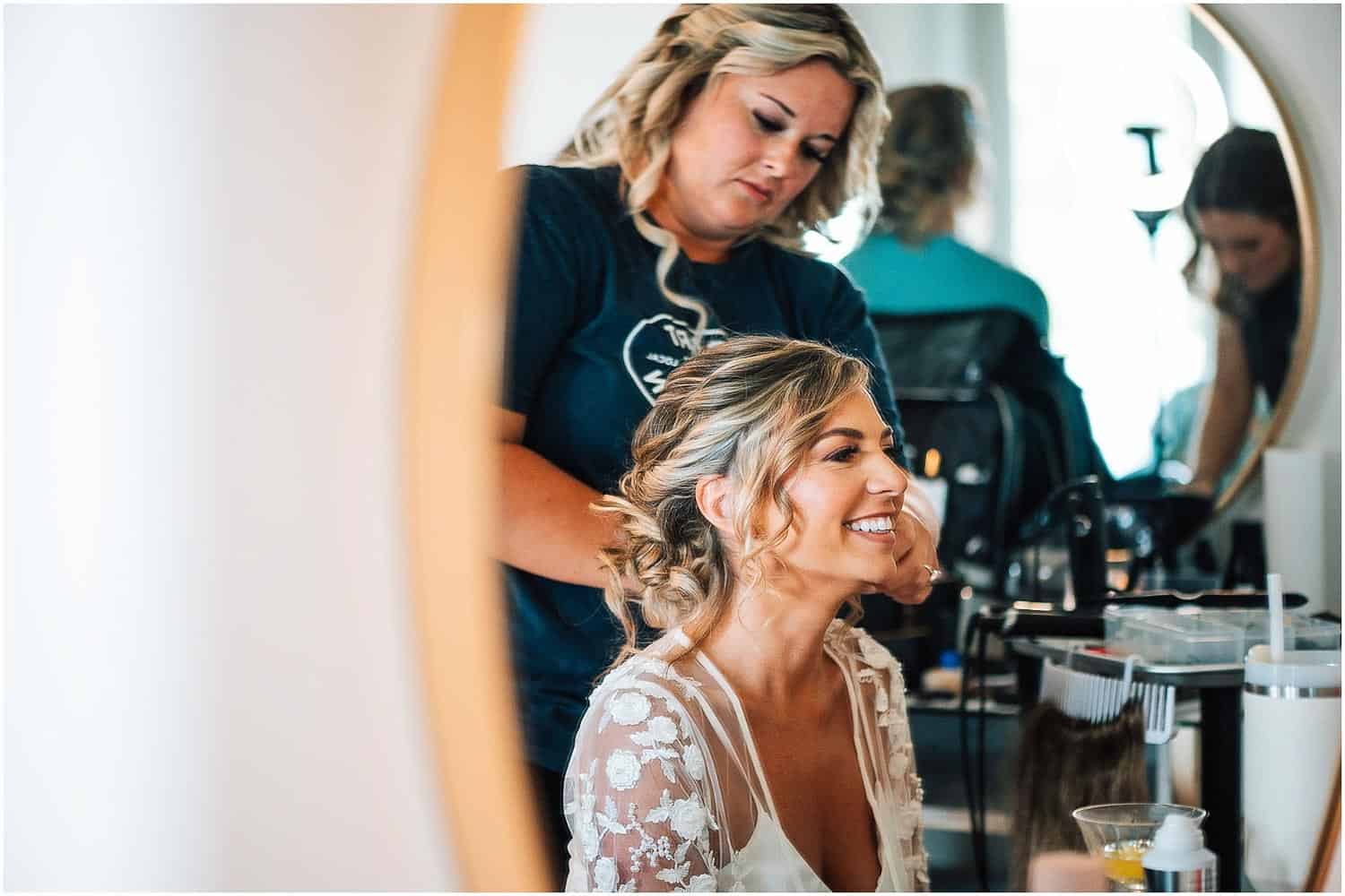 A bride smiling in a salon chair as a stylist fixes her hair, with other people and salon equipment blurred in the background.