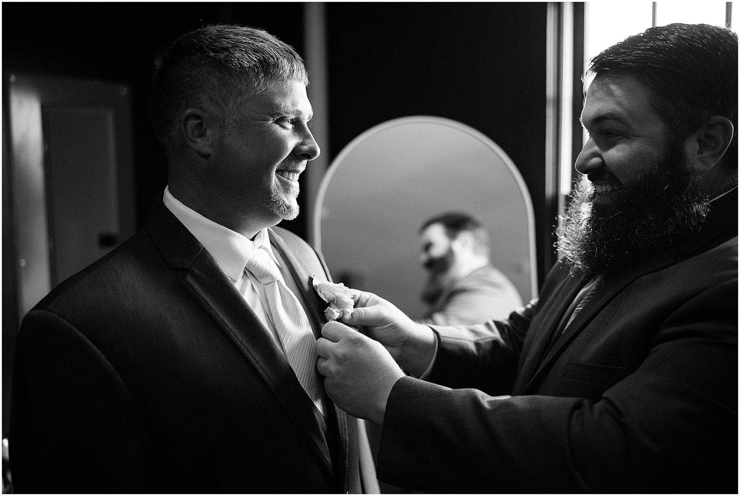 A black and white photo of a bearded man helping another man adjust his tuxedo, both smiling joyfully.