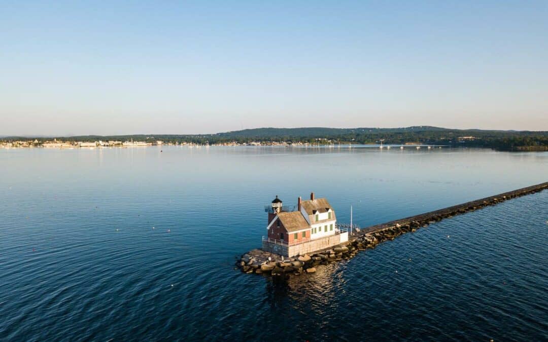 Aerial view of the Rockland Breakwater Lighthouse at the end of a pier, with calm waters and a distant shoreline under a clear sky.