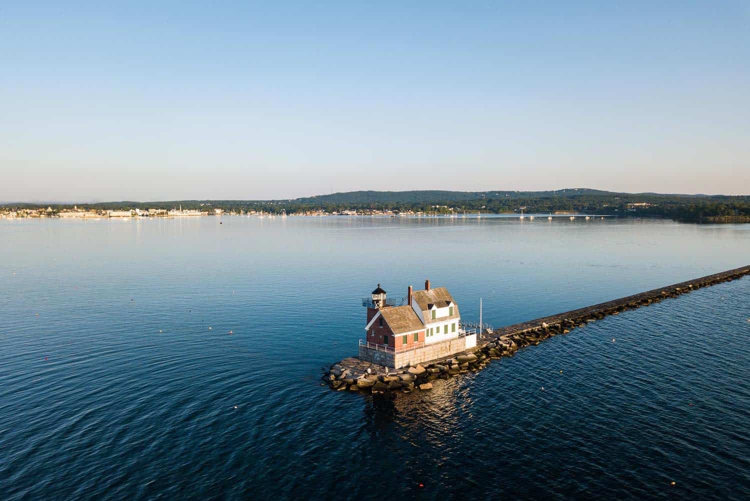 Aerial view of the Rockland Breakwater Lighthouse at the end of a pier, with calm waters and a distant shoreline under a clear sky.