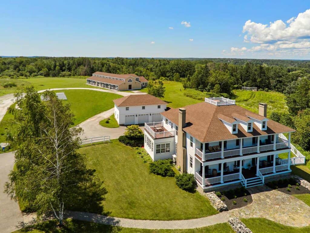 Private estate weddings in maine. Aerial view of ash point estate with multiple residential buildings surrounded by lush green fields under a clear blue sky.