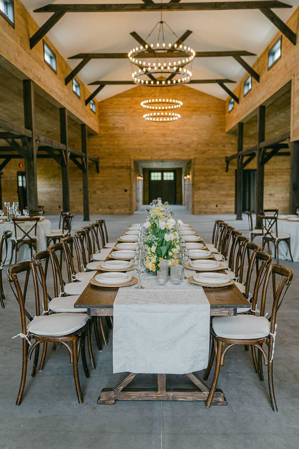 Indoor wedding reception with long tables, white linens, white flower centerpieces, and round chandeliers in a rustic venue.
