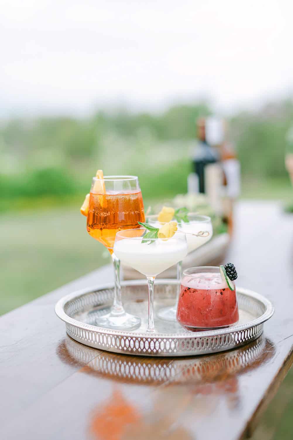 A garnished amber cocktail, a white cocktail with lemon, and a pink cocktail with a berry on a silver tray outdoors.