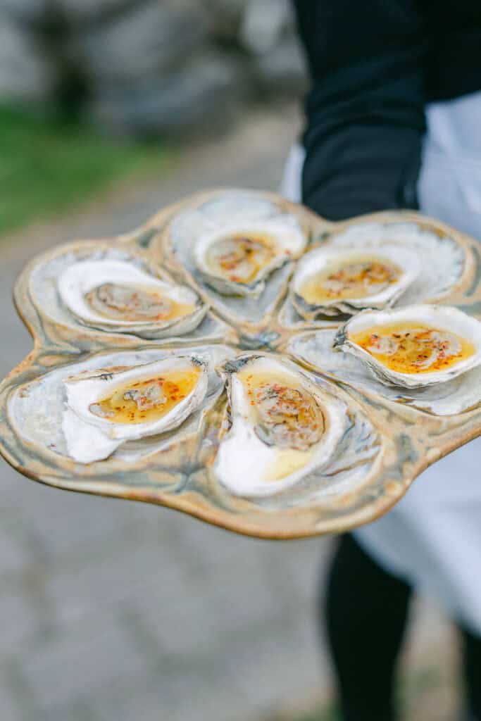 Maine coastal luxury wedding venues, a person holding a plate of six freshly shucked oysters, each topped with a seasoned sauce.